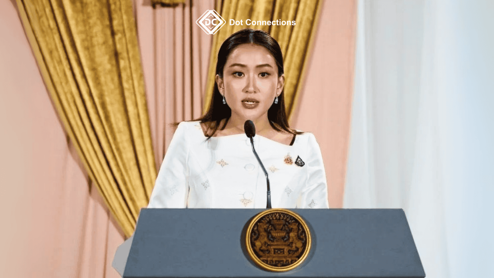A woman in formal white attire speaks at a government podium with the Thai national emblem, standing in front of golden curtains, symbolizing an official government announcement.